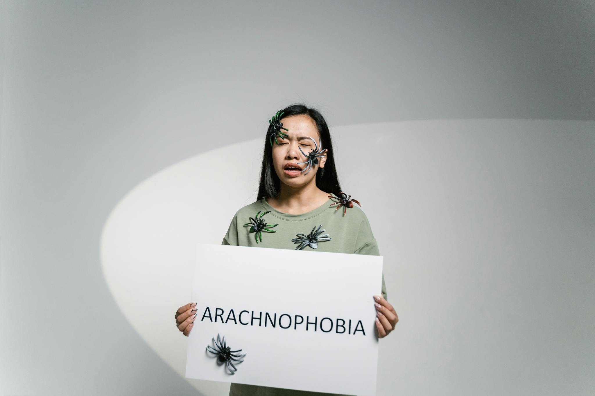 Woman with spiders on face and body holding sign reading 'Arachnophobia'.